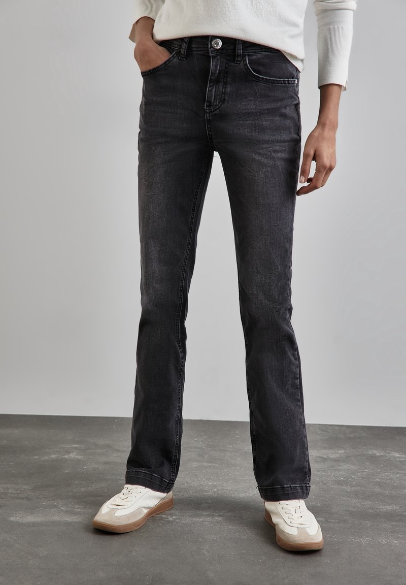 Bas du corps d'une personne portant un jean noir délavé coupe droite, des baskets blanches et un haut blanc à manches longues, debout sur un sol gris.