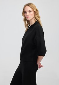 Mladá žena s blond vlasy, oblečená v černé mikině na zip a černých kalhotách, stojící s rukama spojenýma za zády na jednobarevném pozadí.