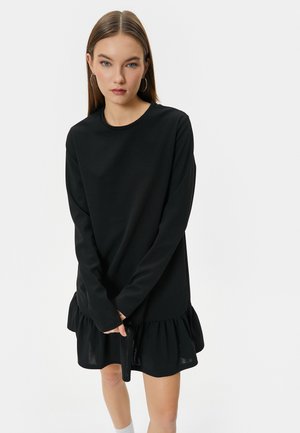 Abito lungo nero a maniche lunghe con scollo tondo e orlo arricciato. Presenta una texture liscia e una vestibilità rilassata. L'orlo si trova sopra le ginocchia.