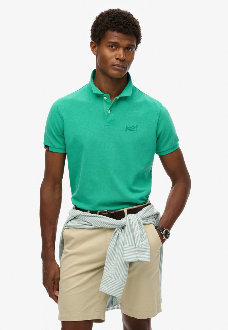 Grøn polo t-shirt med korte ærmer, tekstureret stof, broderet logo på brystet, kombineret med beige shorts og en lyseblå stribet skjorte bundet om taljen.