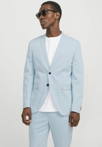 Helles blaues Zweiteiler-Anzug mit einreihigem Blazer und passender Hose. Glatte Textur, schwarze Knöpfe und ein sichtbares, weißes Unterhemd.
