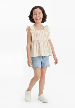 Osmijehnuto dijete s kratkom tamnom kosom koje nosi top bez rukava na pruge s volanima, svijetlo traper kratke hlače i bijele tenisice, stoji na bijeloj pozadini.