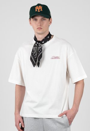 Mladý muž nosí bílé oversized tričko s červeným textem, černý vzorovaný šátek na krku, zelenou baseballovou čepici s oranžovým logem a šedé kalhoty.