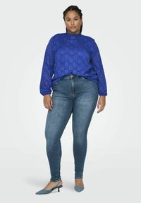 Šviesiai mėlynas ilgomis rankovėmis nėriniuotas marškinėliai su aukšta apykakle ir prigludusia forma, derinami su tamsiai mėlynais džinsais skinny ir šviesiai mėlynais aukštakulniais bateliais.