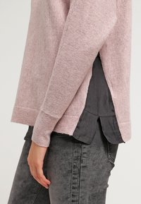 Rosa tröja med en strukturerad yta, med en sidoklyvning och skiktad design över en mörkgrå skjorta. Slim fit, mjukt material.