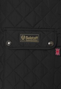 Black quilted jacket featuring a gold "Belstaff" label, snap button closure, and a small Union Jack flag accent. Smooth, matte fabric texture.