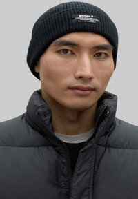 Gorro de canalé negro con una pequeña etiqueta blanca. Lleva puesto con una chaqueta acolchada negra y una camisa gris, mostrando una paleta de colores neutra y monochromática.