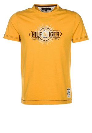Gul t-shirt med korte ærmer med teksten "Hilfiger New York Reg. USA Est. 1985" og et hvidt grafisk patch på den nederste forside.