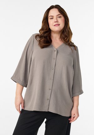 Žena s dlhými vlnitými hnedými vlasmi, oblečená v voľnej taupe blúzke na gombíky s rukávmi dĺžky po lakeť a čiernych nohaviciach, stojaca pred jednoduchým pozadím.