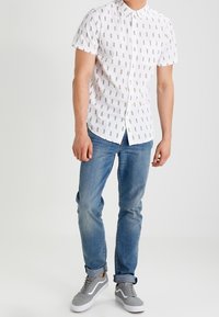 Lyhythihainen valkoinen nappipaita, jossa keltaisia ja harmaita lyijykynäkuvioita, yhdistetty vaaleansinisiin farkkuihin ja harmaisiin lenkkareihin, joissa on valkoisia yksityiskohtia.