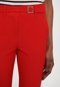 Gros plan d'une personne portant un pantalon rouge vif avec une ceinture assortie et un haut rayé, main reposant à côté de la jambe.
