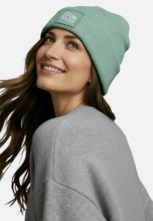 Mint green ribbed beanie with a square camera logo patch, paired with a textured gray sweater. Hair cascades softly over the shoulder.