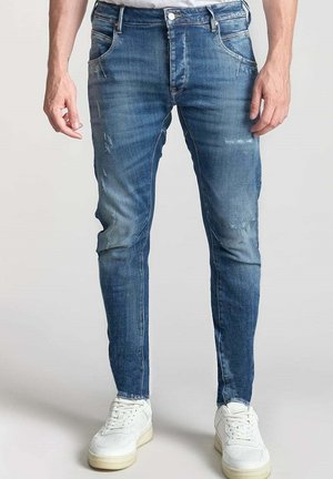 Verbleekte blauwe denim jeans met versleten patches, slim fit, voorzakken en een knoopsluiting, gecombineerd met witte sneakers.