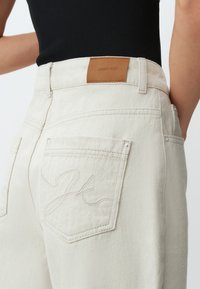 Beige denimbroek met een hoge taille, voorzien van een bruin leren label aan de achterkant en geborduurde details op de rechterzak.