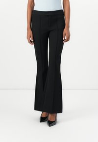 Femme portant un pantalon évasé noir et des talons hauts noirs à bout pointu sur un fond blanc uni.