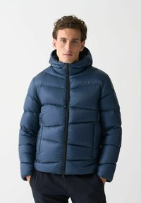 Mann mit lockigem Haar, der eine navyblaue Daunenjacke mit Kapuze trägt, die Hände in den Taschen, steht vor einem schlichten hellen Hintergrund.
