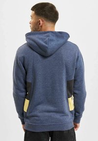 Sudadera con capucha azul de acabado texturizado, con acentos en bloques de color negro y amarillo en la parte baja de la espalda y los lados.