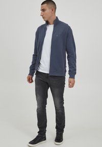 Chaqueta de hombre de color azul marino con cremallera, hecha de tela suave, que presenta un cuello alto, puños de canalé y un diseño ajustado, combinada con jeans grises y zapatillas negras.