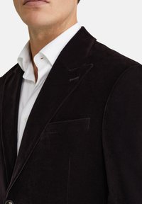 Tumšs samta žakets ar vienu krūšu kabatu, izgrieztiem atloks un gludu tekstūru, apvienots ar tīru baltu krekls zem tā.