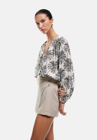 Langärmlige Bluse in Creme mit schwarzem Blumenmuster, ausgestattet mit einem V-Ausschnitt und gefältelten Ärmeln, kombiniert mit beigen High-Waist-Shorts.