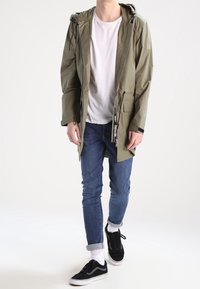 Une personne portant une veste à capuche vert olive, un t-shirt blanc, un jean bleu retroussé, des chaussettes blanches et des baskets noires marchant en avant.