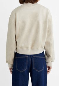 Beige ruskindsjakke med ribbet halsudskæring og manchetter, afslappet pasform. Har syningsdetaljer langs ryggen. Kombineret med mørkeblå denimjeans.