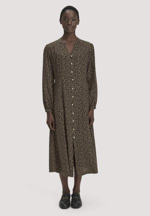 Vrouw draagt een lange bruine jurk met witte polkadots, voor geknoopt, lange mouwen en zwarte loafers, staand tegen een effen achtergrond.