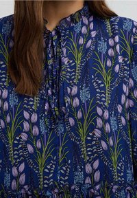 Blå blus med lila och grönt blommönster, med en rynkad krage och en knuten rosett vid halsringningen.