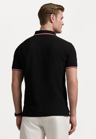 Homem de costas, vestindo uma camisa polo preta com detalhes em vermelho e branco na gola e nas mangas, combinada com calças brancas.