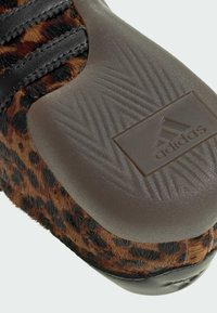 Tenisky s leopardím vzorom, s textúrovanou hnedou gumovou podrážkou, čiernymi textilnými popruhmi a embosovaným logom Adidas na podrážke.