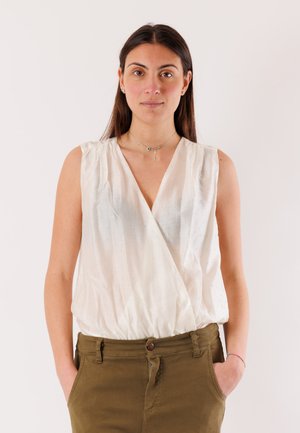 Donna con capelli lisci e scuri che indossa una camicetta bianca senza maniche e pantaloni verde oliva, in piedi con le mani nelle tasche davanti a uno sfondo semplice.