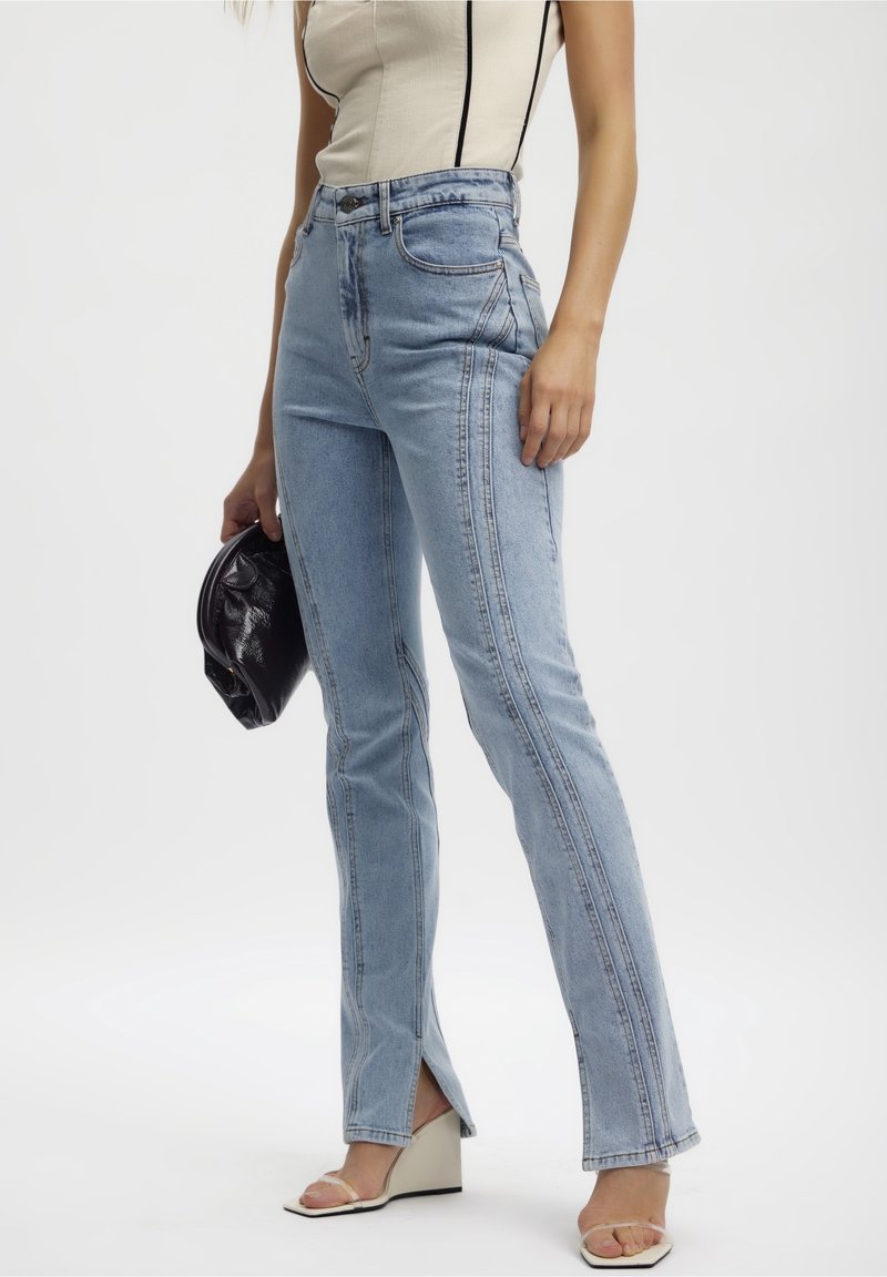 Vaqueros de tiro alto en azul claro con costuras verticales, leve acampanado en la parte inferior y aberturas laterales. Combinados con un bolso negro y sandalias beige.