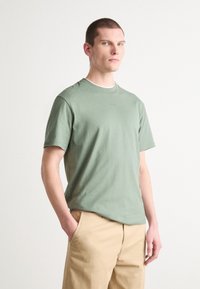 T-shirt en coton vert clair avec des manches courtes, un col rond et un logo discret au centre. Associé à un pantalon beige.
