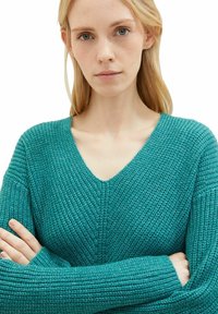 Türkis gestrickter Pullover mit V-Ausschnitt, strukturiertem Stoff und langen Ärmeln, der einen lässigen Schnitt und ein sichtbares Rippenmuster aufweist.
