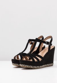 Černé wedge sandály s více páskovým designem, zsuede materiálu a jute obloženou platformou. Nastavitelný pásek kolem kotníku a hladký interiér.