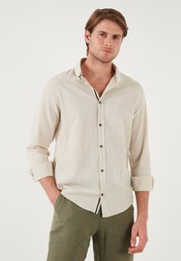 Camisa de lino beige claro con un diseño abotonado, mangas enrolladas y una textura sutil. Presenta acentos a rayas contrastantes en el interior del collar.