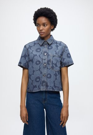 Γυναίκα με κοντά κατσαρά μαλλιά που φοράει κοντομάνικο, κουμπωτό τζιν πουκάμισο με σκοτεινά λουλουδάτα σχέδια και σκούρα τζιν ψηλόμεσα.