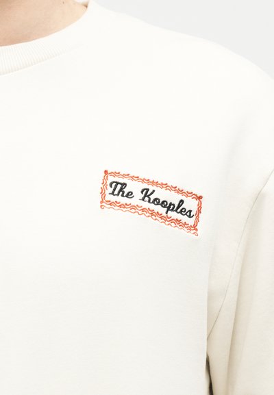 Λευκό πουλόβερ με στρογγυλή λαιμόκοψη και το κέντημα "The Kooples" στο στήθος, μέσα σε ένα πορτοκαλί ορθογώνιο με κυματιστές άκρες.