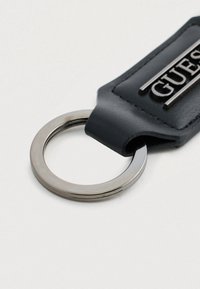 Portachiavi in pelle nera con anello circolare in argento e una targhetta rettangolare incisa con la scritta "GUESS" in caratteri metallici.