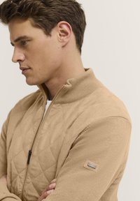 Beige gesteppte Jacke mit hohem Kragen, Vorderreißverschluss und gerippten Bündchen. Verfügt über ein kleines Etikett am linken Arm. Glatte Textur.