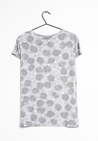 Szary T-shirt z krótkim rękawem wiszący na czarnym wieszaku, z wzorem różnej wielkości szarych kółek na jaśniejszym szarym tle.