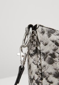 Bord de sac à main texturé noir et blanc à motif serpent, avec un clip en métal argenté et une attache de sangle noire. Surface lisse avec coutures visibles.