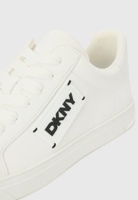 Hvit sneaker med en glatt syntetisk overdel, flate snører, og svart "DKNY"-merke i et hevet rektangulært skilt på siden.