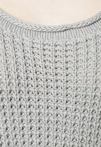 Gros plan sur un tissu tricoté gris clair avec un motif côtelé vertical et un col arrondi sur un fond de peau claire.