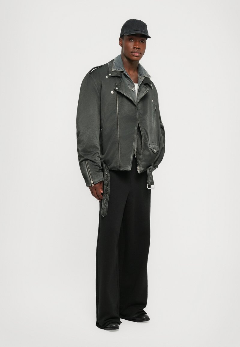 Gray faux leather motorcycle jacket with silver zippers, side pockets, and adjustable straps. Paired with black wide-leg trousers and a cap.