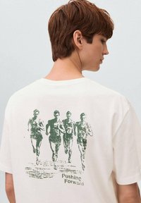 Ung person med kort brunt hår iført en hvid t-shirt med grøn grafik af fire løbende atleter og teksten "Pushing Forward" på ryggen.