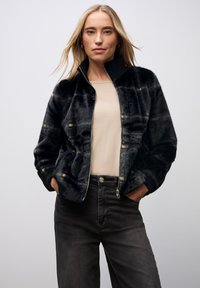 Sort faux-fur bomberjakke med lynlås foran og ribbet krave, prydet med et subtilt rutemønster og sidelommer, kombineret med grå jeans.
