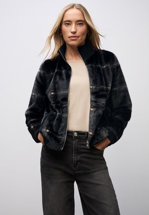 Chaqueta bomber de pelo sintético negra con cierre de cremallera y cuello de canalé, que presenta un sutil patrón de cuadros y bolsillos laterales, combinada con jeans grises.