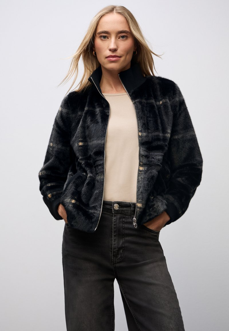Sort faux-fur bomberjakke med lynlås foran og ribbet krave, prydet med et subtilt rutemønster og sidelommer, kombineret med grå jeans.