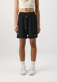 Black cotton shorts with elastic waistband and drawstring, featuring side pockets and printed text detail on leg. Worn with white sneakers and socks.